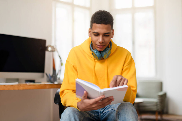 A young man in a yellow hoodie reads from a notebook, seated comfortably at a home office space - Photo, Image