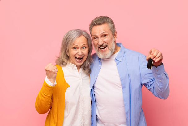 Joyful casal de cabelos grisalhos seniores segurando as chaves de seu novo carro e gesticulando sim, comemorando sua conquista da propriedade do automóvel contra o pano de fundo do estúdio rosa. Transporte, auto empréstimo - Foto, Imagem