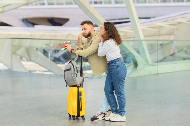 Αναστατωμένο νεαρό ζευγάρι έχασε την πτήση ή το τρένο, και οι δύο στέκονται δίπλα σε μια φωτεινή κίτρινη βαλίτσα σε ένα σύγχρονο σταθμό διέλευσης - Φωτογραφία, εικόνα