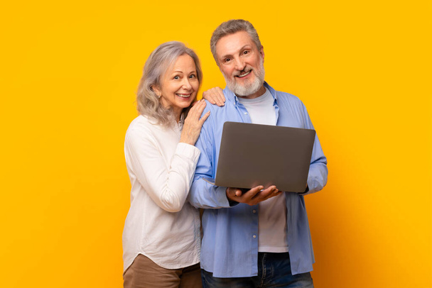 モダンなラップトップでオンラインブラウジングを楽しむシニアカップル, 黄色の背景の上にコンピュータデバイスと立って, 高齢者のスタジオショットは,彼らの退職で技術の容易さを紹介しています - 写真・画像