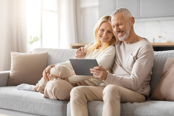 Loving mature couple sharing a tablet and smiling while seated comfortably on their couch at home - Photo, Image