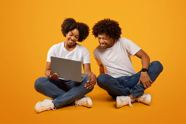 Pretty young black woman showing her husband something on laptop. Happy loving african american couple sitting on floor and using computer, checking educational website, yellow background - Fotó, kép