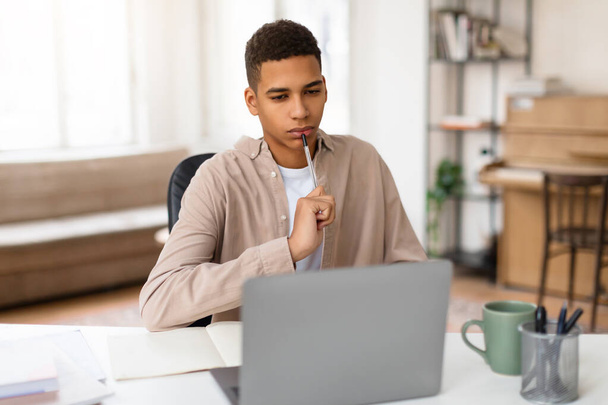 Adolescente negro contemplativo con la pluma a la barbilla, enfocado intensamente en la pantalla de su computadora portátil mientras trabaja en un proyecto educativo o aprendizaje a distancia en casa - Foto, imagen