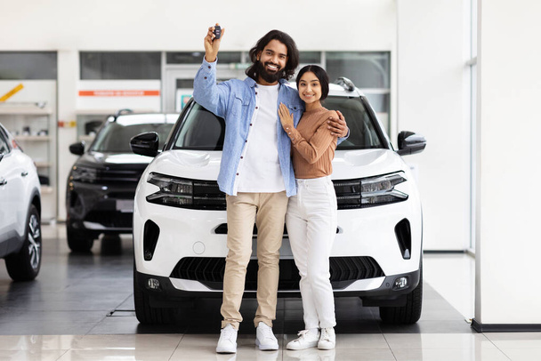Feliz pareja oriental abrazando y sosteniendo una llave de coche delante de un nuevo vehículo blanco en un concesionario de automóviles - Foto, imagen