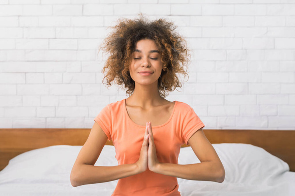 Afrikanisch-amerikanische Frau sitzt in namaste Yoga-Position und entspannt sich morgens auf dem Bett - Foto, Bild