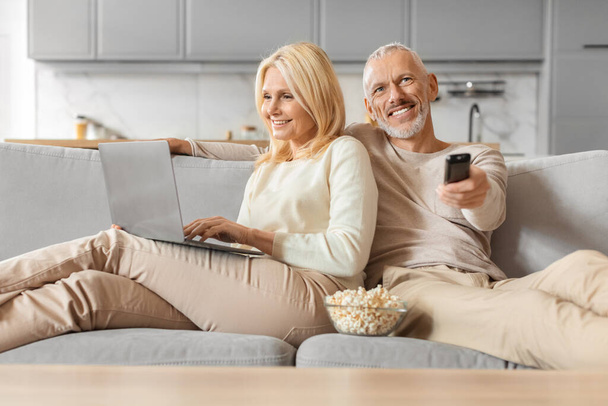 Couple sénior multi-tâches avec ordinateur portable et télécommande, passer une soirée détendue à la maison - Photo, image