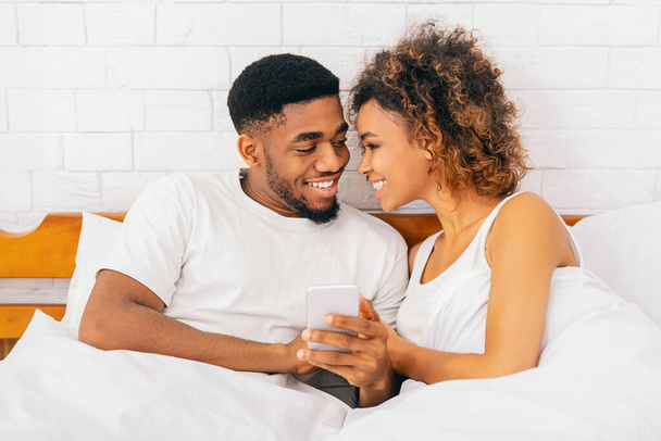 Между нами нет секретов. Счастливые афро-американец мужчина и женщина читают электронную почту по телефону, улыбаясь друг другу в постели
 - Фото, изображение