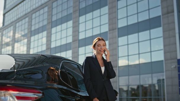 笑顔の女性ボスは,豪華な車の上に傾いている携帯電話を呼び出します. オフィスビルの近くにスマートフォンを呼ぶ美しいリラックスしたビジネスマン. 幸せな女性 ceo は電話で成功した交渉を持っています - 写真・画像