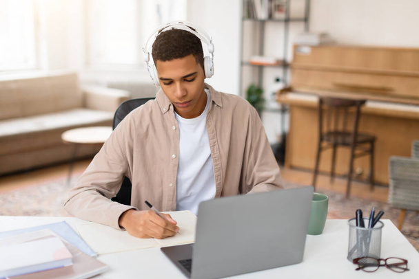 Hombre joven enfocado con auriculares blancos escribe en un cuaderno con su portátil abierto, comprometido en el aprendizaje - Foto, imagen