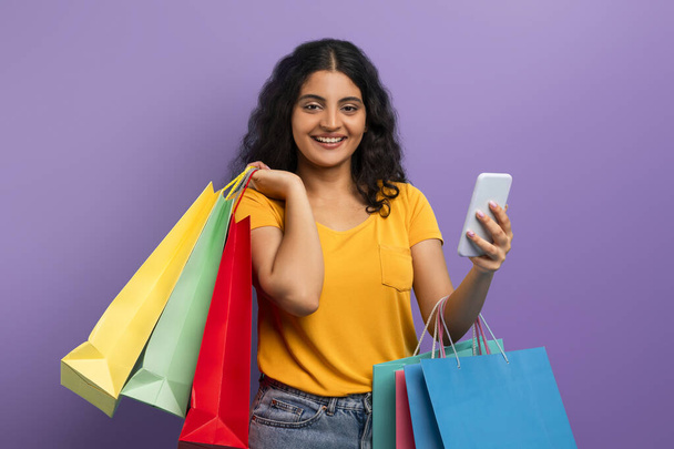Een vrouw in een gele top glimlacht met boodschappentassen en een smartphone, wat een positieve winkelervaring suggereert - Foto, afbeelding