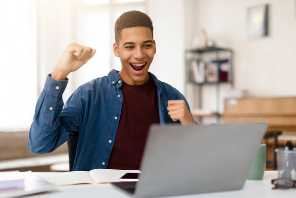 Uitbundige zwarte tiener kerel juicht met vuist pomp terwijl hij kijkt naar zijn laptop, teken van triomf en opwinding in het bereiken van persoonlijk doel of overwinning - Foto, afbeelding