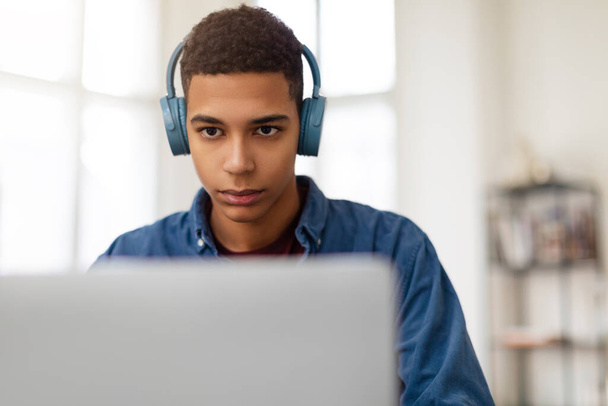 Keskittynyt nuori mies, jolla on siniset kuulokkeet, toimii kannettavalla tietokoneella, katsoen tarkkaavaisesti näyttöä - Valokuva, kuva