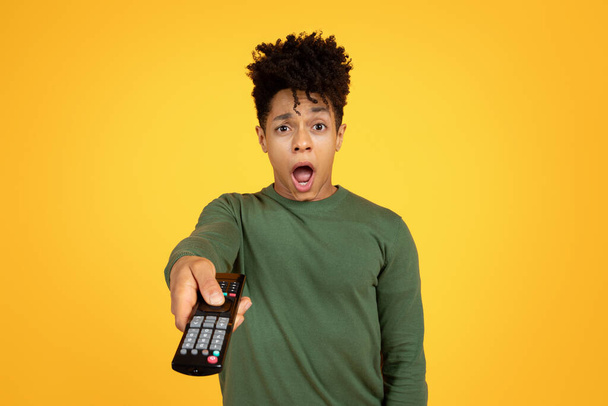 Un hombre negro sorprendido sosteniendo un mando a distancia y expresando shock sobre un fondo amarillo - Foto, imagen