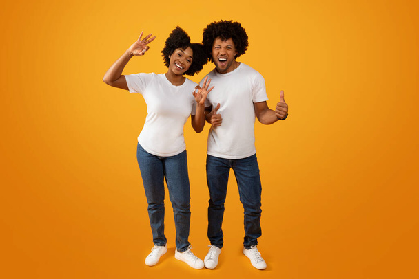 Cool african american millennial man and woman with bushy hair in casual gesturing isolated on yellow studio background. Happy black couple showing okay and thumb up, full length - Zdjęcie, obraz