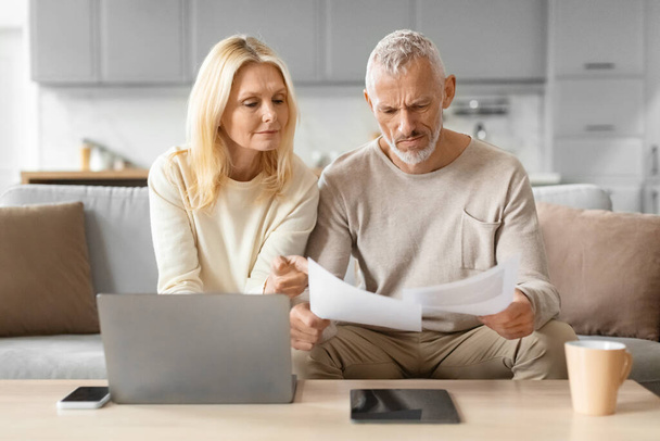 Elderly couple analyzing papers with attention, using a laptop and tablet for reference at home - Foto, Imagen