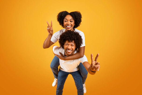 Cool pretty millennial african american woman with bushy hair piggybacking her boyfriend or husband, happy black couple gesturing showing peace and smiling, isolated on yellow background - Фото, изображение