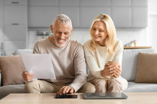 Ένα μεσήλικα ζευγάρι κάθεται στον καναπέ, κοιτάζοντας ένα έγγραφο χαρτί, χαμογελώντας, σε ένα άνετο περιβάλλον στο σπίτι - Φωτογραφία, εικόνα