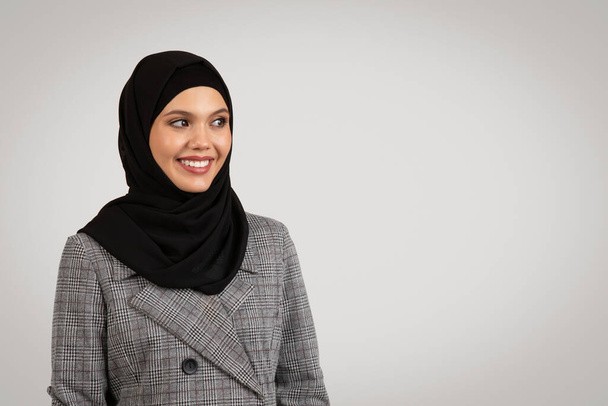 Magabiztos és vidám professzionális üzletasszony hidzsábot és houndstooth öltönyt visel, oldalról nézve a szabad térben, fénysemleges háttérrel - Fotó, kép