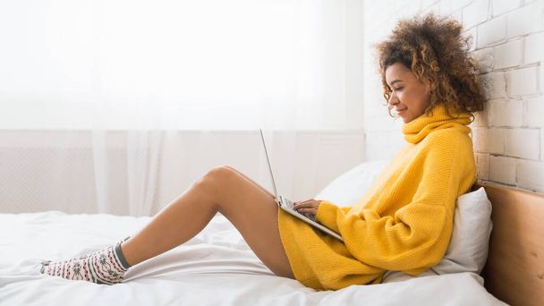 Mujer afroamericana trabajando en la computadora portátil, sentada en la cama en casa, espacio para copiar, vista lateral
 - Foto, imagen