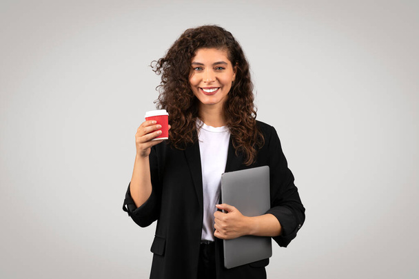 Elinde kahve fincanı ve gri bir dizüstü bilgisayarı olan kendine güvenen genç bir kadın gri bir arka plana karşı poz veriyor, profesyonelliği ve ulaşılabilirliği tasvir ediyor. - Fotoğraf, Görsel