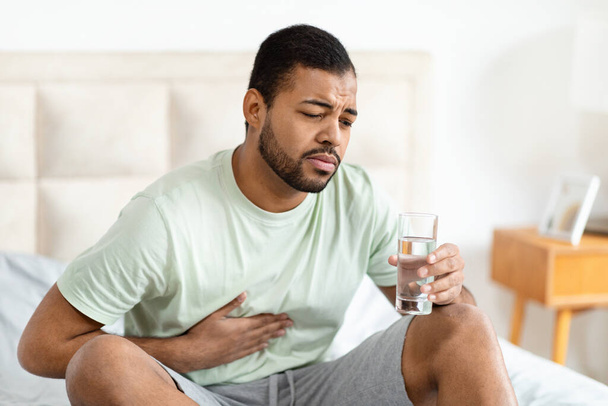 Αφροαμερικάνος κάθεται στο κρεβάτι κρατώντας το στομάχι του και ένα ποτήρι νερό, απεικονίζοντας δυσφορία ή ασθένεια σε ένα οικιακό περιβάλλον - Φωτογραφία, εικόνα