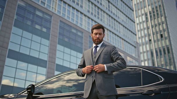Rich businessman posing at car buttoning jacket zoom out. Elegant bearded man millionaire standing urban street near automobile premium class. Confident wealthy ceo relaxing outdoors feeling success. - Фото, изображение