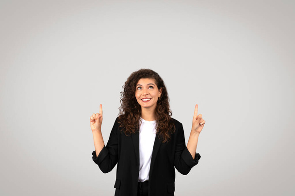 Veselá žena s kudrnatými vlasy ukazuje oběma prsty vzhůru, stojí na prostém pozadí, vyzařuje pozitivitu a sebedůvěru - Fotografie, Obrázek