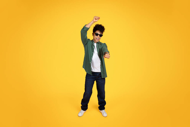 Energetico giovane afroamericano in abbigliamento casual che balla gioiosamente, catturato in una posa a metà movimento su uno sfondo giallo - Foto, immagini