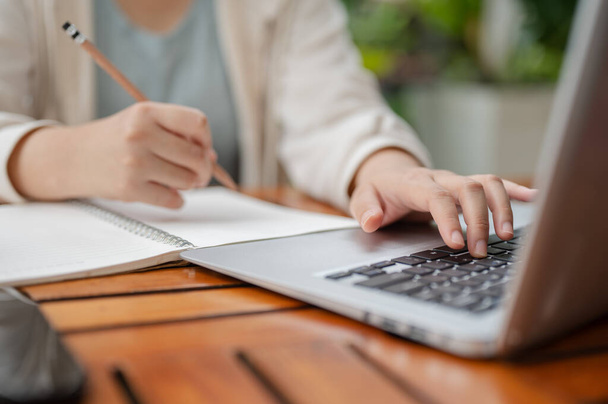 Uma imagem recortada de uma mulher em desgaste casual está digitando no teclado do laptop e escrevendo ou tomando notas no notebook enquanto trabalha remotamente em um espaço ao ar livre. - Foto, Imagem