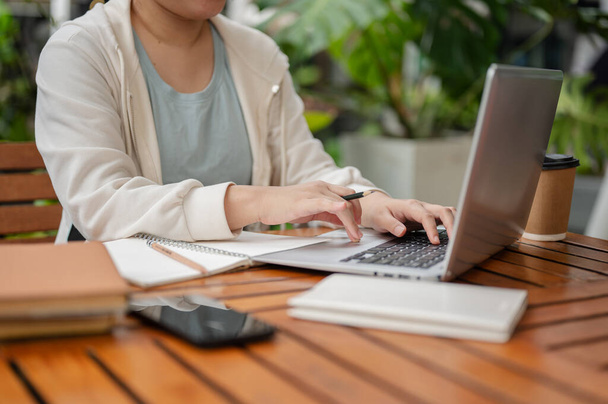 A cropped image of an Asian woman in casual wear is holding a pencil while typing on laptop keyboard, working remotely at an outdoor space. - Photo, Image