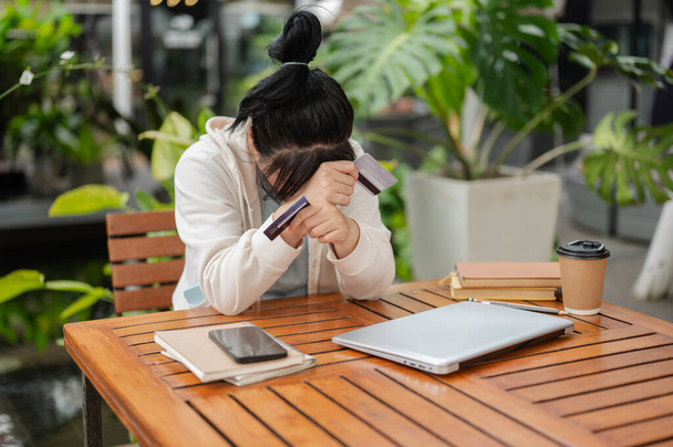A stressed, upset Asian woman sitting at an outdoor cafe table, hand covering her face while holding a credit card, having financial problems or bankrupt. - Photo, Image