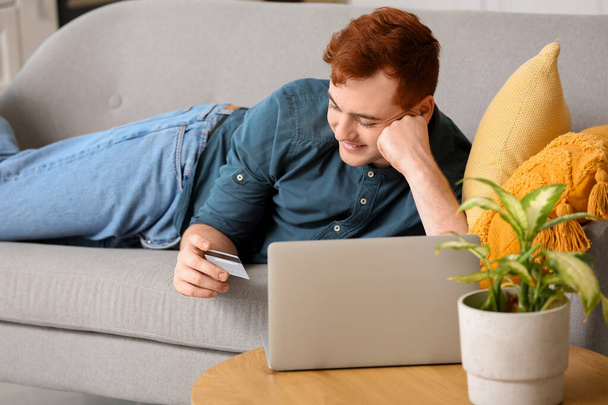 Beau jeune homme avec carte de crédit et ordinateur portable moderne couché sur le canapé tout en faisant des achats en ligne - Photo, image