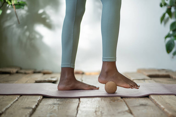 Beine rollen Fitnessball auf Yogamatte. Afroamerikanerin Selbstpflege-Training mit myofaszialem Massageball, rollenden Füßen, Entspannung und Verbesserung der Durchblutung. Sportgeräte für einen gesunden Lebensstil. - Foto, Bild