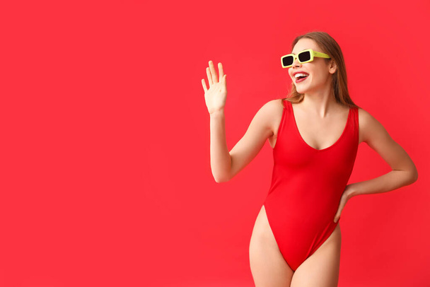 Beautiful young woman in tank swimsuit waving hand on red background - Photo, Image