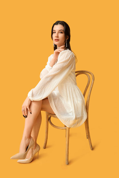 Piękna młoda kobieta ze stylową biżuterią siedzi na krześle na żółtym tle - Zdjęcie, obraz