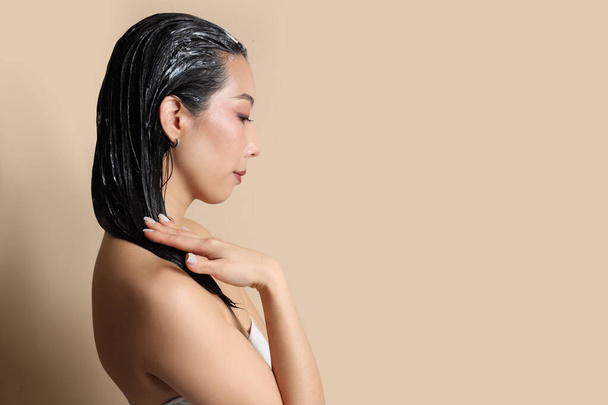 Mujer asiática joven aplicando mascarilla para el cabello sobre fondo beige, primer plano - Foto, imagen