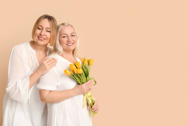 Adult woman with her smiling mother and bouquet of tulips on beige background. International Women's Day celebration - Photo, Image