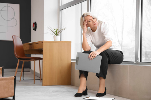 Madura mujer de negocios con portapapeles experimentar la menopausia en el alféizar de la ventana en la oficina - Foto, Imagen