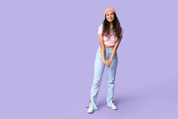 Cool African-American woman wearing top, jeans and hat on lilac background - Photo, Image