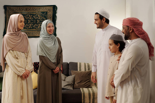Moderni muslimi miehet, naiset ja lapsi yllään vaatimaton vaatteita seisoo olohuoneessa ottaa keskustelun perheen kerääminen Eid Al-Fitr - Valokuva, kuva