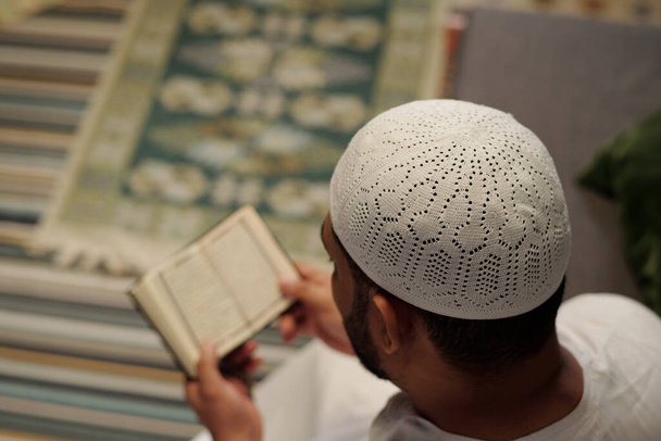 Wysoki kąt widzenia selektywne ujęcie ostrości nierozpoznawalnego muzułmanina w białym taqiyah czytającego Koran, przestrzeń kopiowania - Zdjęcie, obraz