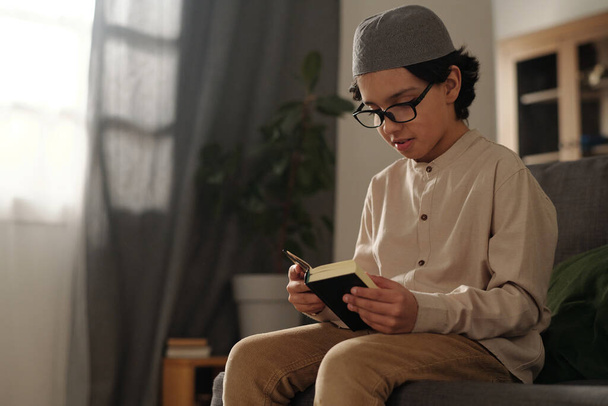 Mediana toma de niño musulmán con anteojos sentados en el sofá leyendo libro sagrado del Corán, espacio para copiar - Foto, imagen