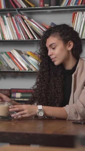Kobieta z kręconymi włosami siedząca w bibliotece, mieszająca kawę i pijąca. średni strzał - pionowy - Materiał filmowy, wideo