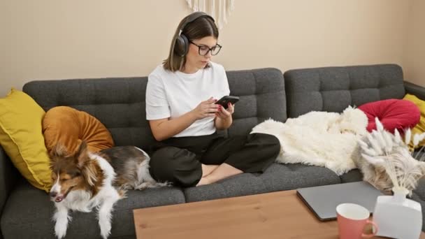 Una donna contenta con le cuffie gode della musica su uno smartphone mentre si rilassa su un divano a casa con i suoi cani dormienti. - Filmati, video