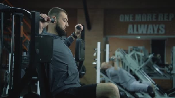 Motivasyonlu atletik adam protez bacaklı, spor aletleriyle modern fitness merkezinde egzersiz makinesine bağlanmış. Arka planda başka bir yetişkin sporcu kol egzersizi yapıyor.. - Video, Çekim