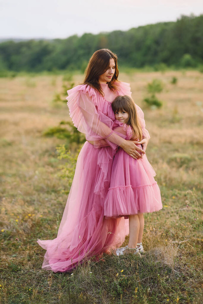 Mother and her daughter in tulle pink dresses  hugging on nature at summer. Beautiful mummy and her little girl in same designed long pink dresses. Love. Family concept. Photoshooting - Photo, Image