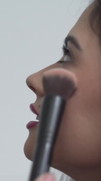 Hermosa dama colombiana mirando hacia arriba y sonriendo mientras el artista del maquillaje aplica rubor en su mejilla. primer plano, vista lateral - Vertical 1080 - Imágenes, Vídeo