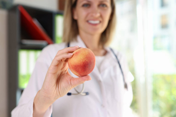Smiling female nutritionist holds fresh peach fruit in her hand close-up. Doctors advice, recommendations and diet for healthy lifestyle. - Photo, Image