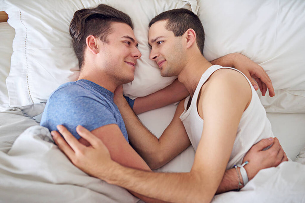 Lächeln, Männer und Blick von oben auf ein schwules Paar zu Hause im Bett, um sich morgens zu entspannen, zu umarmen und miteinander zu verbinden. Lgbtq, oben und glückliche Menschen im Schlafzimmer für Liebe, Verbindung und gesunde Beziehung. - Foto, Bild