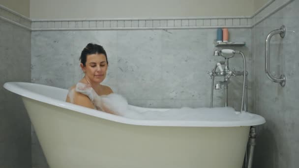 リラックスした若い女性は,モダンな家庭内インテリアでヴィンテージの爪の足の浴槽でバブル浴を楽しんでいます. - 映像、動画